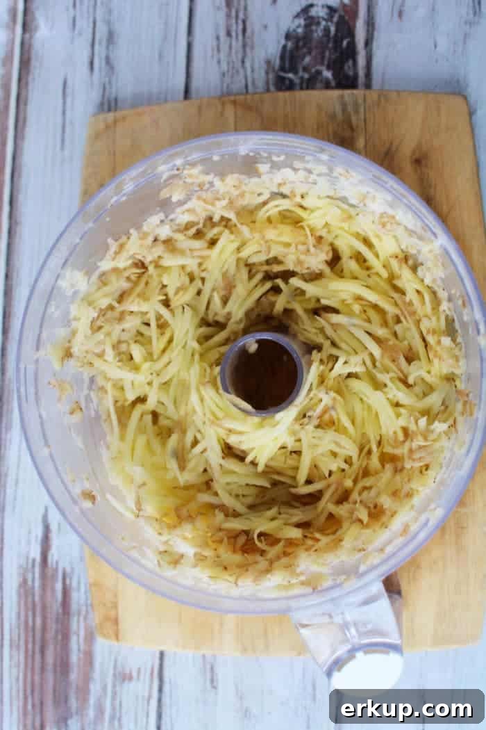 Shredded potatoes in a food processor bowl on a wooden cutting board, ready to be transformed into crispy Air Fryer Latkes.