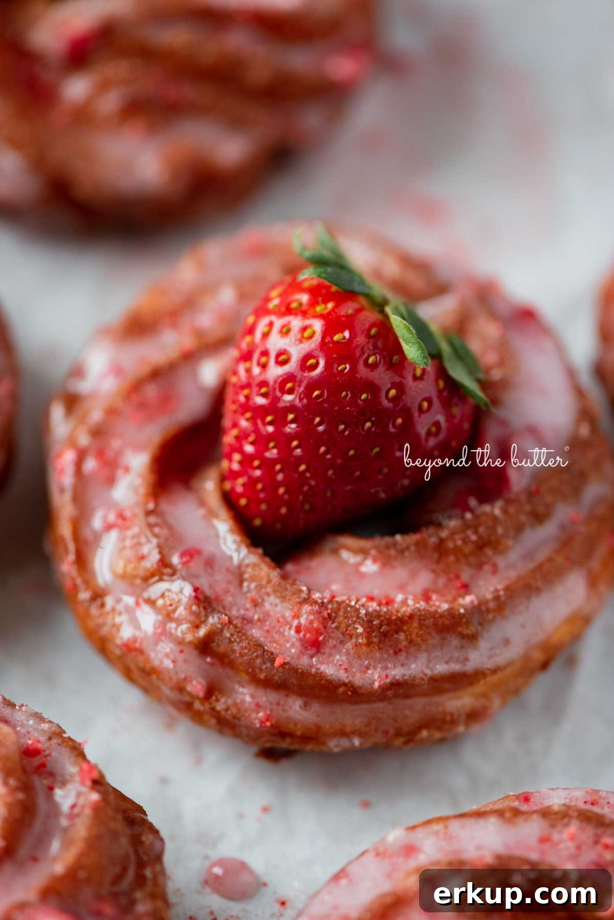 Just fried strawberry glazed french crullers on a parchment paper lined baking sheet topped with a fresh strawberry