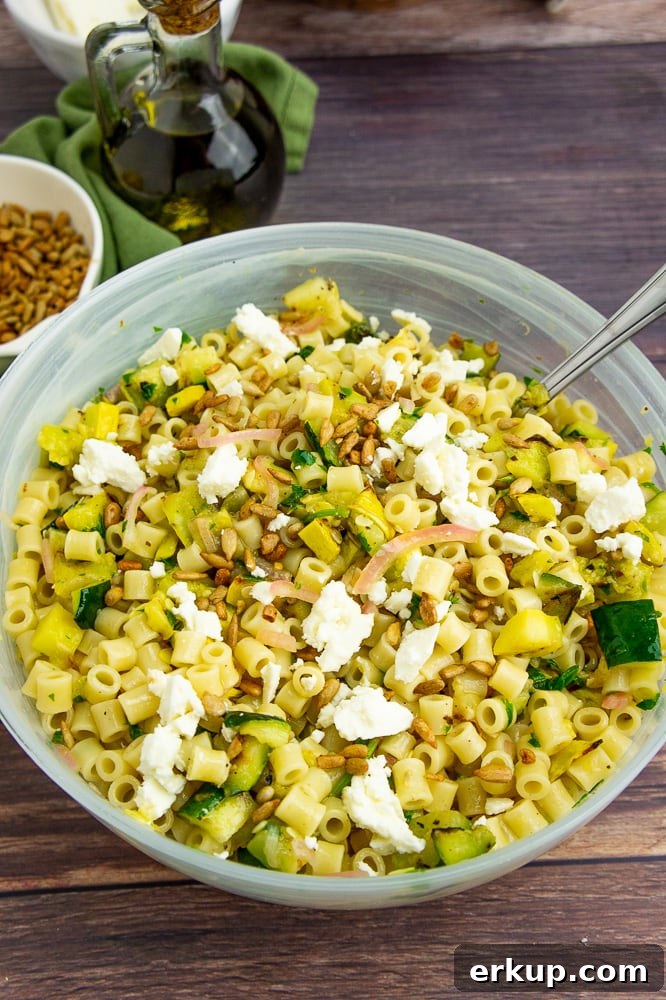 a bowl of pasta salad with a silver serving spoon topped with crumbled white cheese and sunflower seeds on a wooden table.
