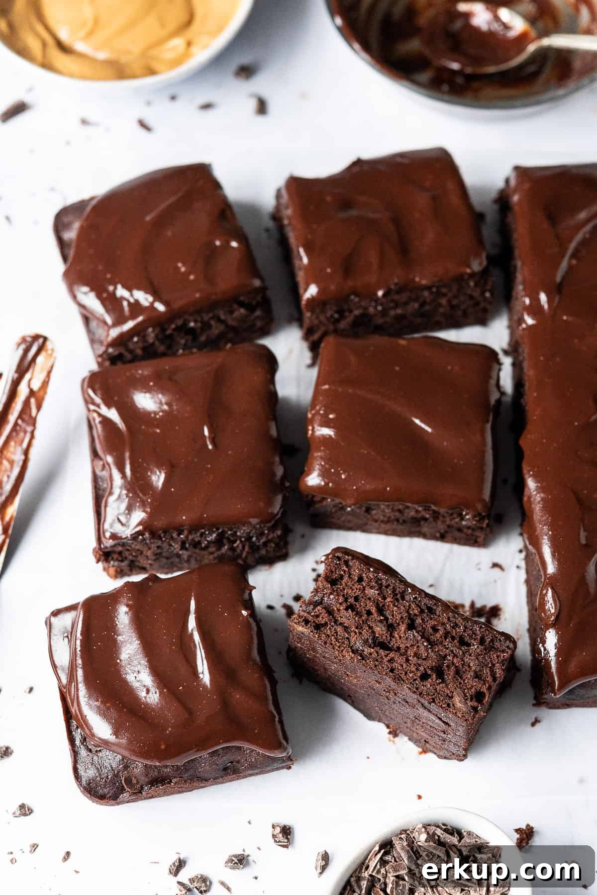 Squares of vegan chocolate banana cake on white baking parchment with a bowl of peanut butter and bowl of ganache in the background.