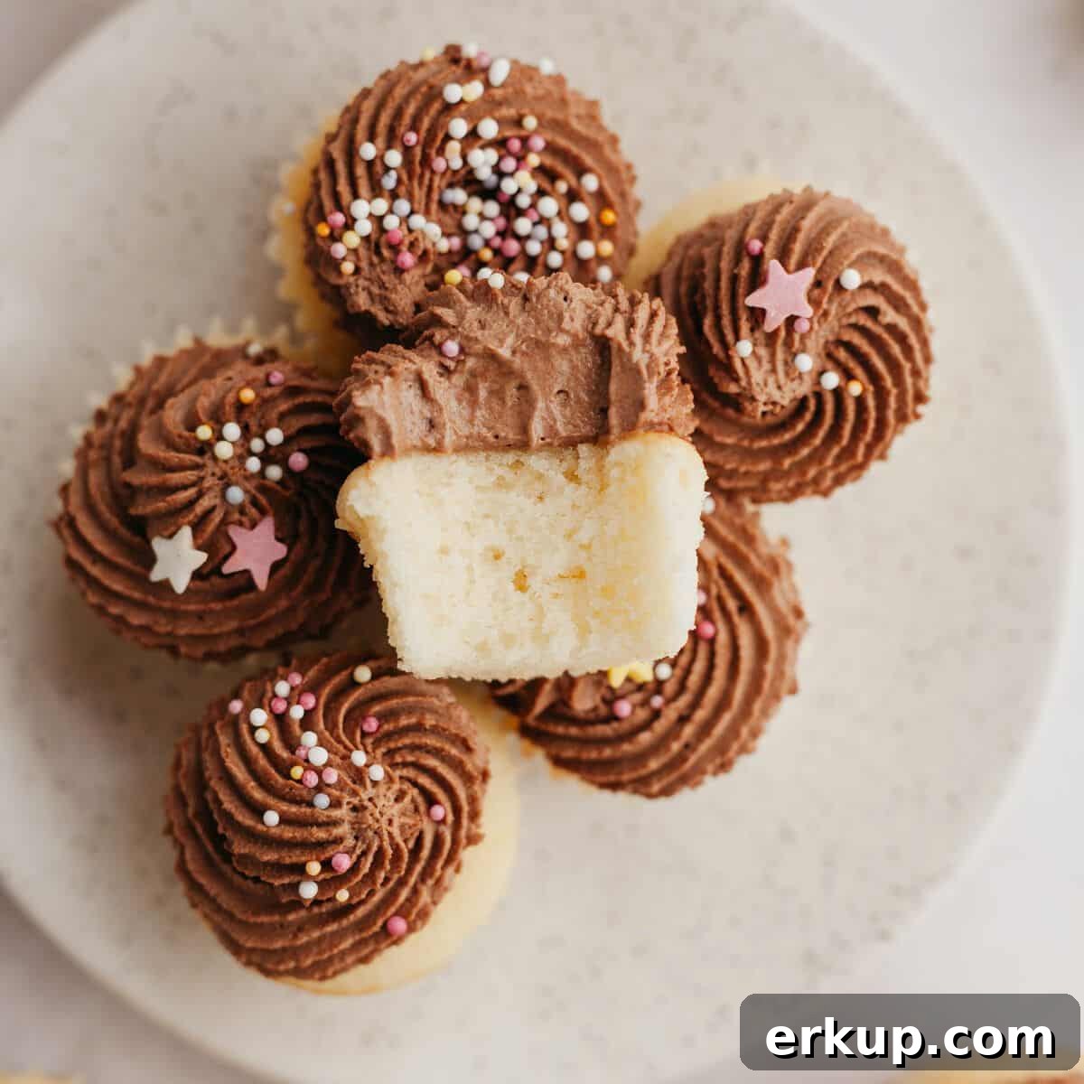 A bunch of mini vanilla cupcakes with chocolate frosting on a plate, one cupcake is on top with a bite taken out of it.
