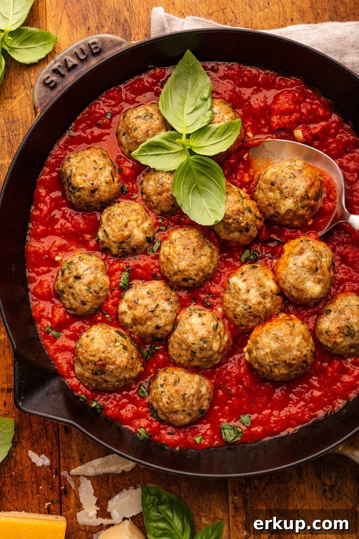 Classic Italian Chicken Meatballs in Marinara Sauce 2 A skillet pan with red marinara sauce and baked Italian chicken meatballs in it. The pan of meatballs is displayed on a wooden background with fresh basil leaves garnishing the dish and placed around the pan along with a light gray napkin and shredded cheese.
