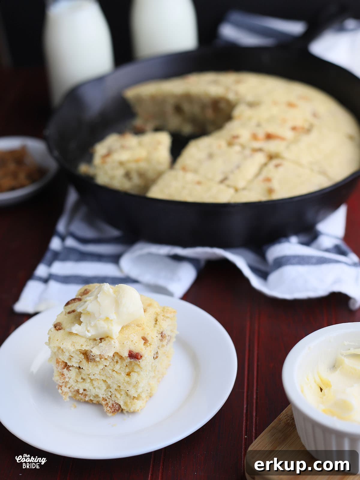 side shot of a slice of cracklin' corn bread topped with a dollop of butter on a white plate with a cast iron skillet of bread in the background
