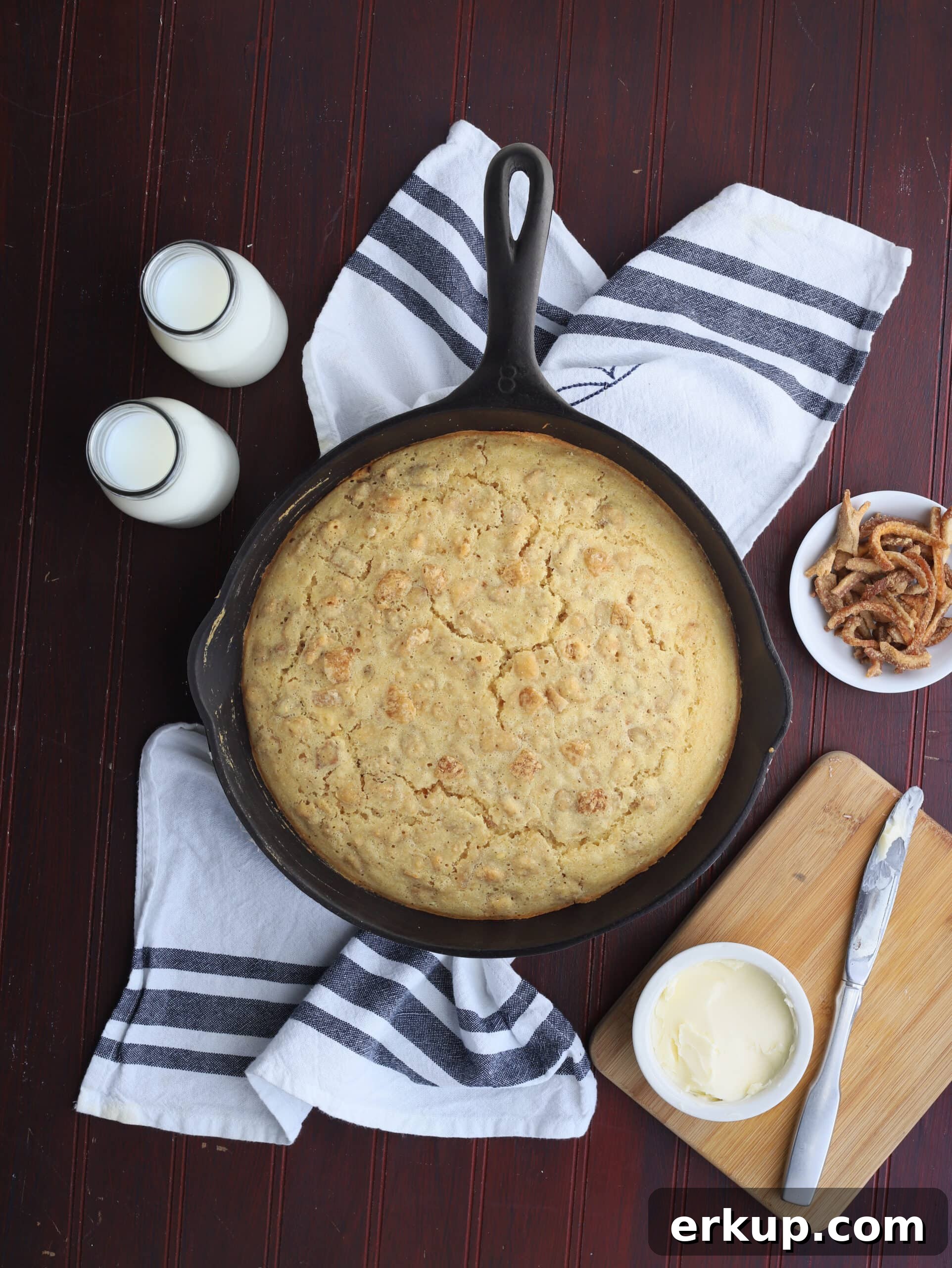 overhead shot of cracklin' corn bread in a cast iron skillet with butter, milk and a dish or cracklings to the side