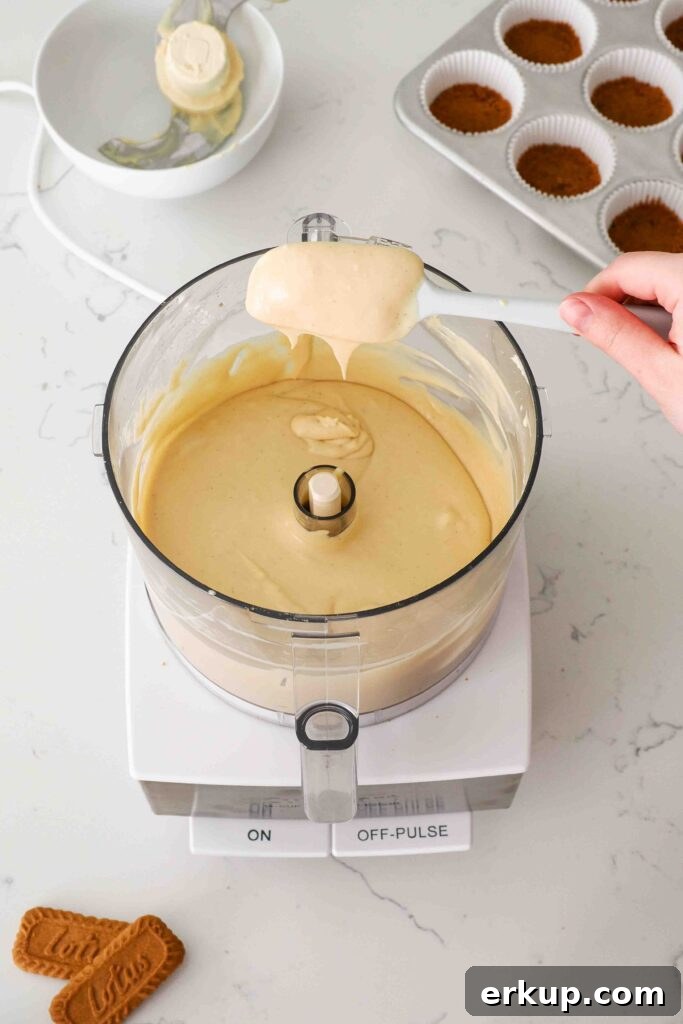 A spatula holds up Biscoff cheesecake batter over a food processor bowl.