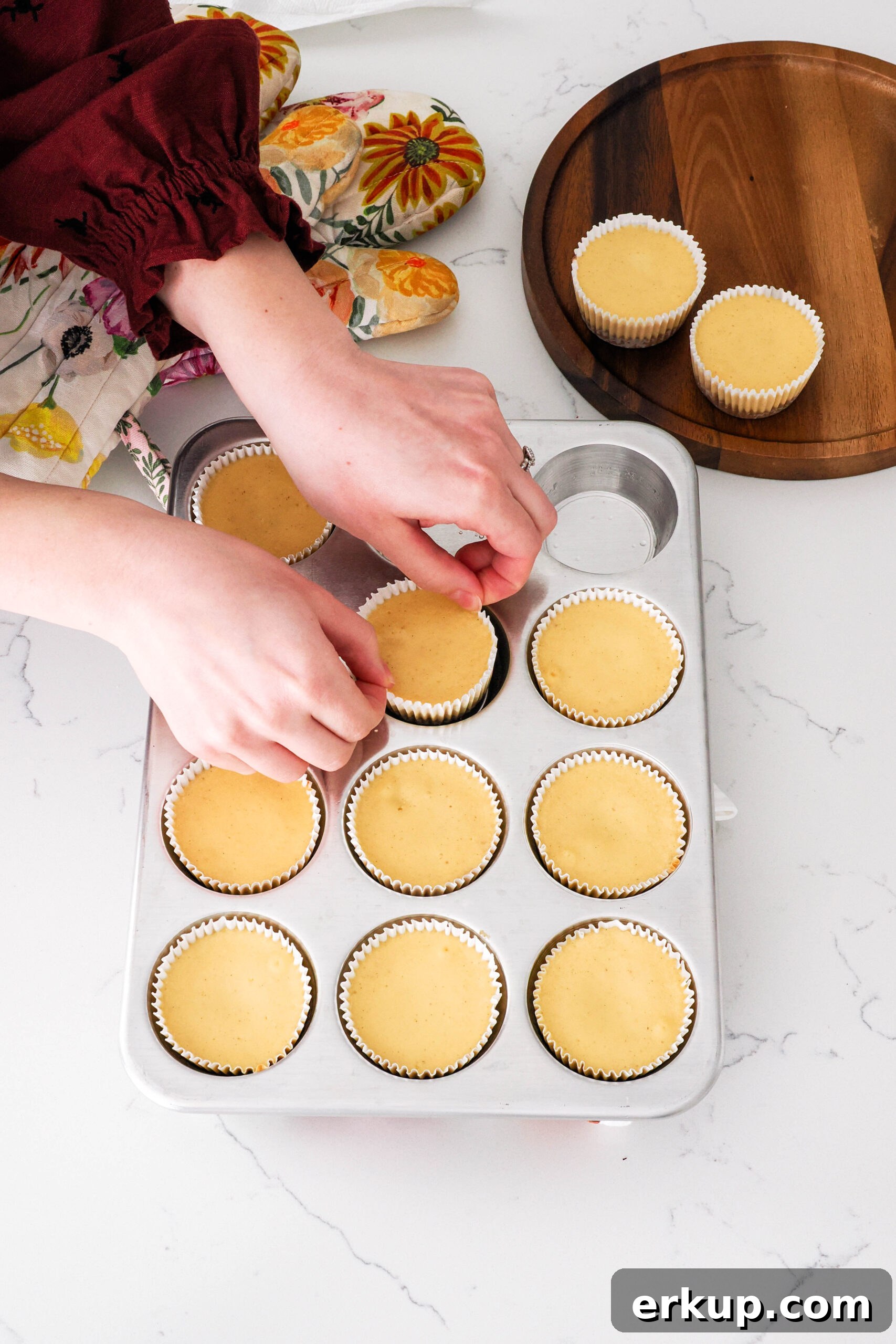 Two hands pinch the edges of the paper liner on a mini cheesecake to remove it from a muffin pan.