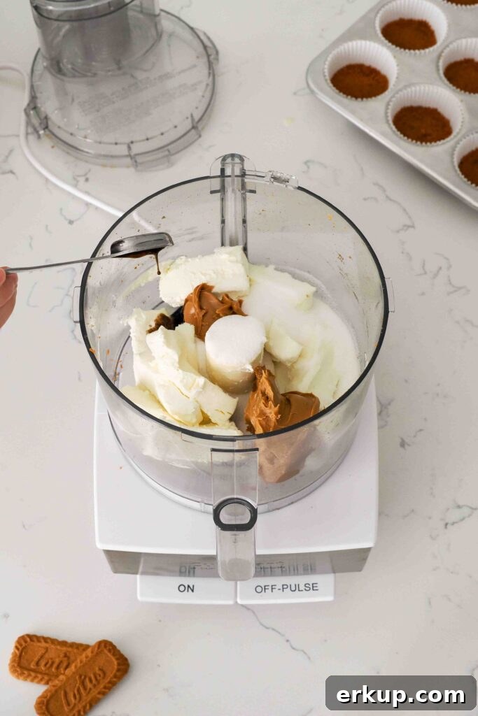 A hand adds vanilla bean paste to a food processor bowl with cream cheese, cookie butter, and sugar.