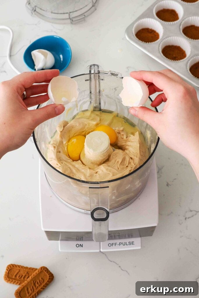 Two hands break an egg into the bowl of a food processor with Biscoff cheesecake filling.