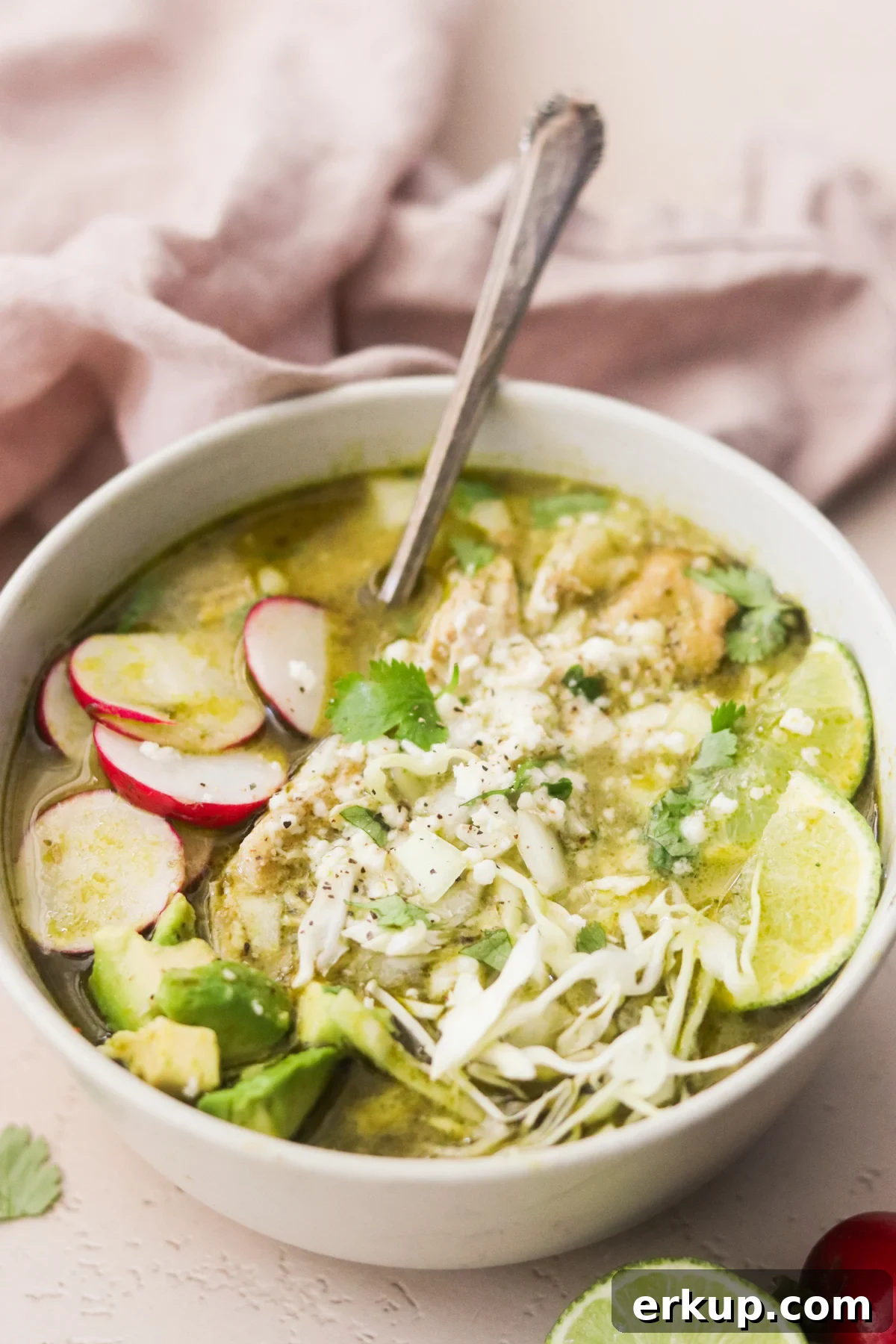 Bowl of pozole verde with tender pork, shredded cabbage, radish, avocado, and lime wedges.