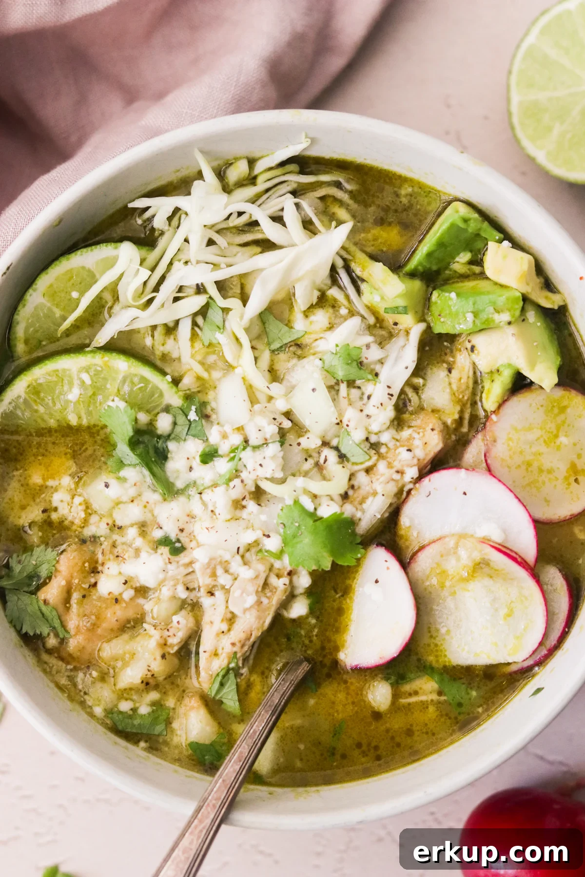 Overhead view of a spoon resting in a bowl of pozole verde topped with avocado, radish, cabbage, and cilantro.