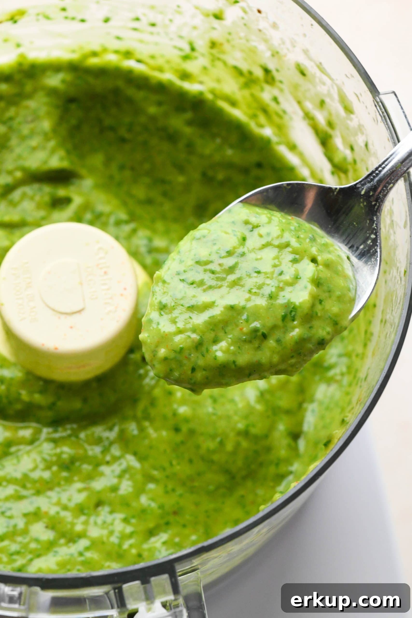 How to make avocado herb green sauce: A spoon lifting out a small amount of sauce from the food processor to show the creamy texture.
