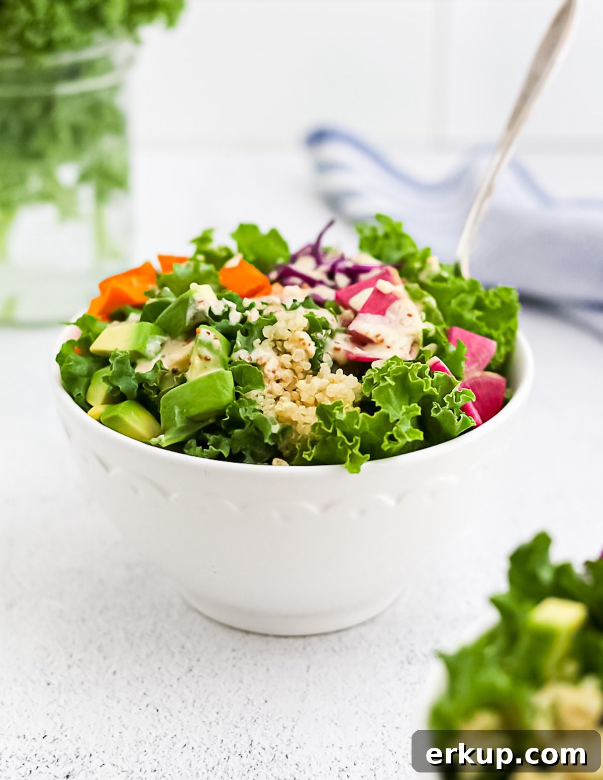 A kale salad with tahini dressing in a white bowl, containing radishes, sweet potatoes, cabbage, and avocado.