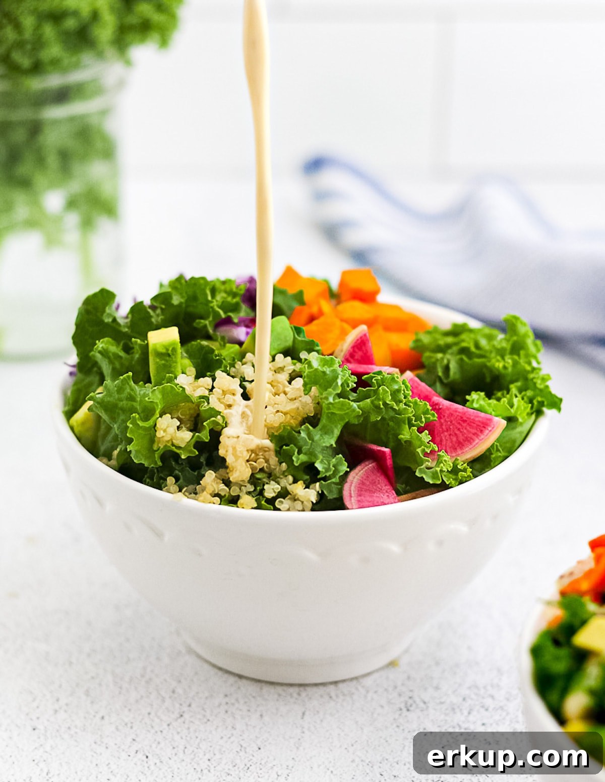 A picture of tahini salad dressing being poured onto a salad with kale, radishes, sweet potato, avocado, and cabbage.