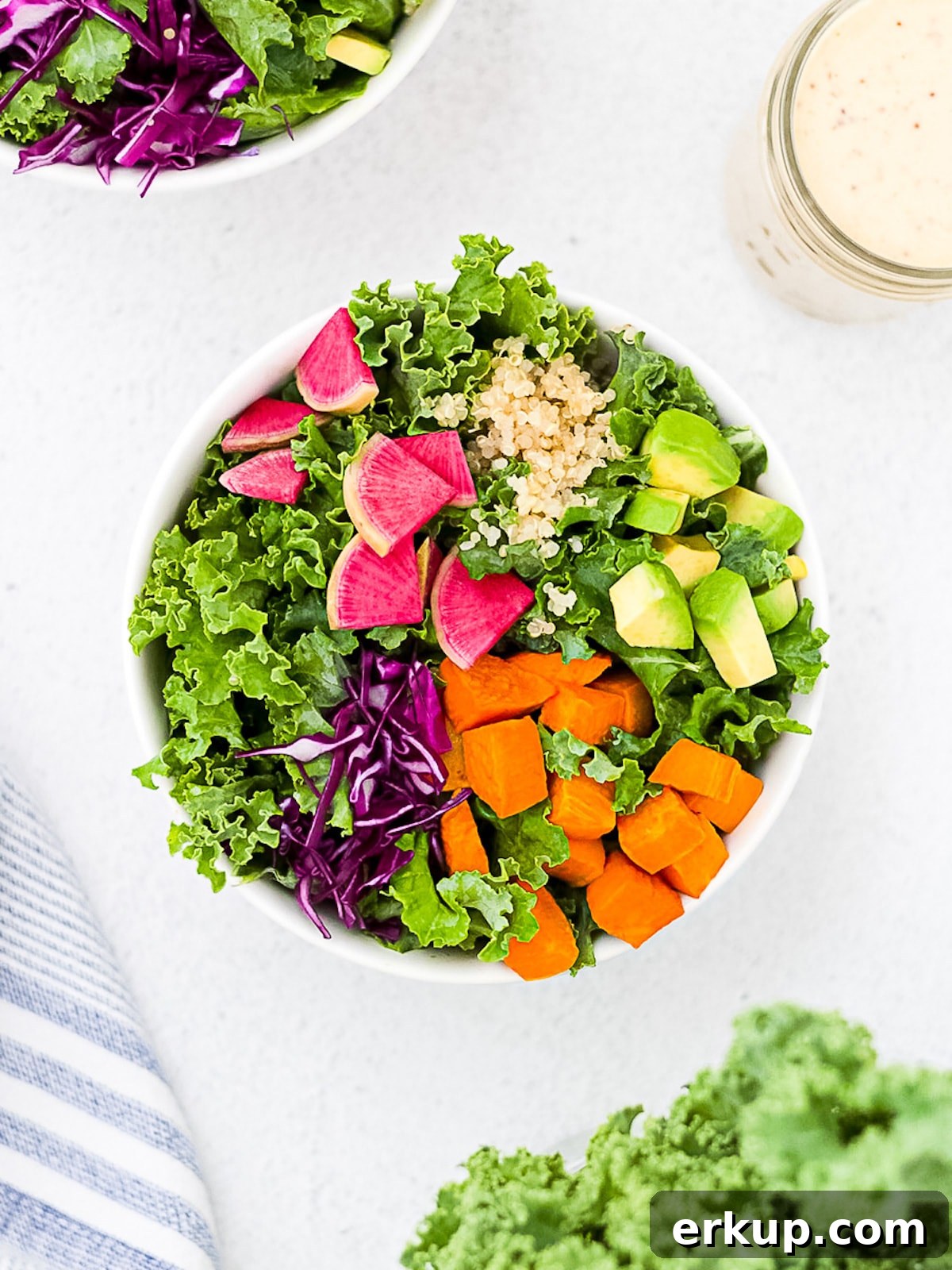 A white bowl filled with prepared kale, pink radishes, sliced avocado, roasted and diced orange sweet potatoes, sliced red cabbage, and cooked quinoa.