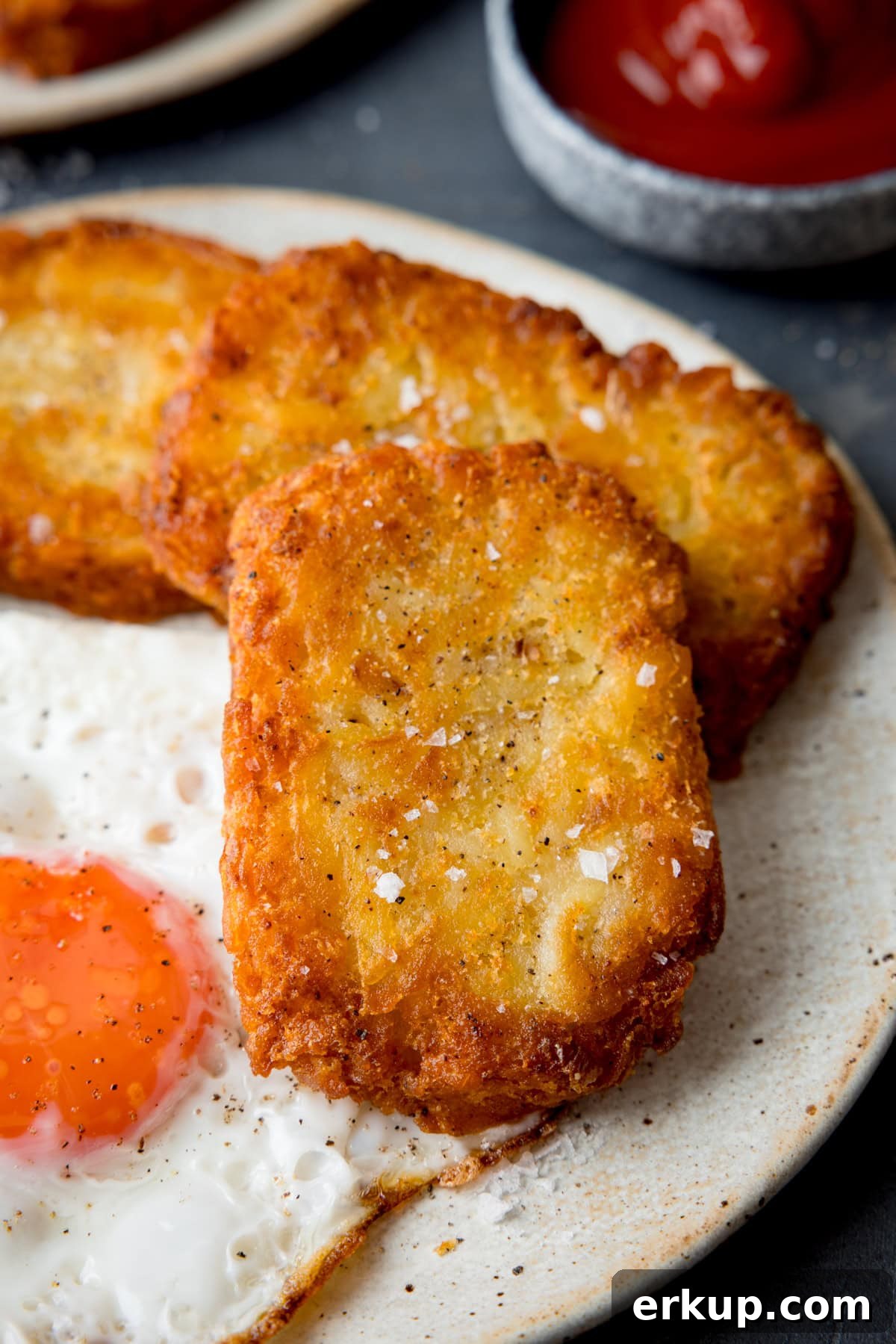Crispy Homemade Hash Browns Recipe 2 A tall, close-up shot of three hash browns on the side of a large white plate, which has a fried egg in the centre. In the top left of the image, you can slightly see a second white plate. In the top right-hand side of the image, there is a dark blue dish filled with ketchup. This is all set on a navy blue background.