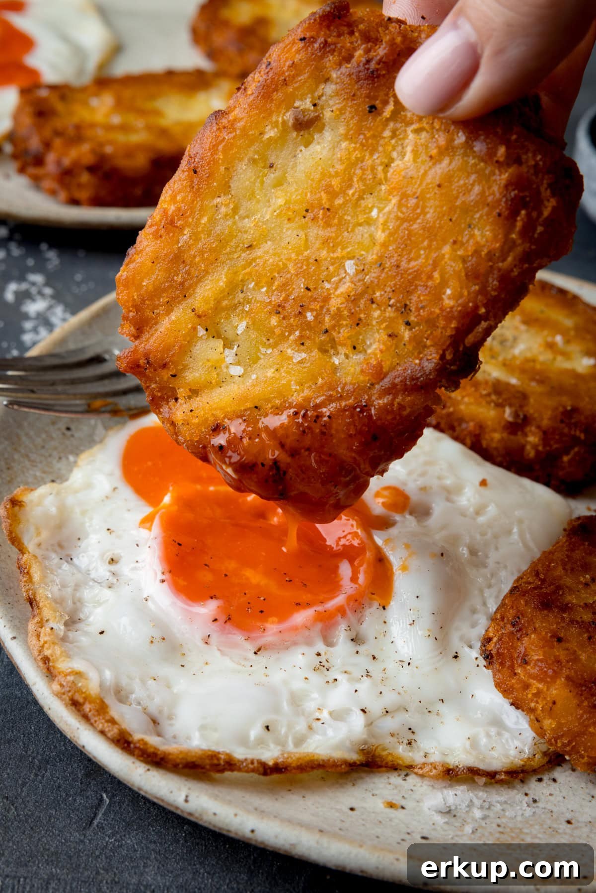 Crispy Homemade Hash Browns Recipe 5 A tall, close-up shot of three hash browns on the side of a large white plate, which has a fried egg in the centre, and a silver fork set at an angle on the left-hand side of the plate. In the centre of the image, there is one whole hash brown being held by a hand coming from the top centre of the image, the hash brown is being dipped into the yolk of the egg. In the top left of the image, you can slightly see a second white plate with two hash browns and a fried egg on it. This is all set on a navy blue background.