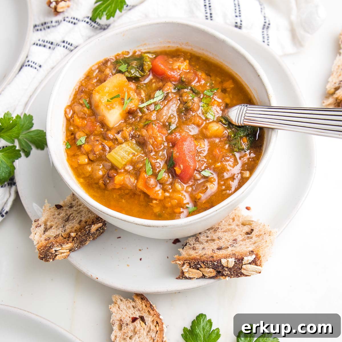 Chunky lentil and vegetable soup with crusty chunks of bread, fresh sprigs of parsley and a white with navy striped dish towel. 