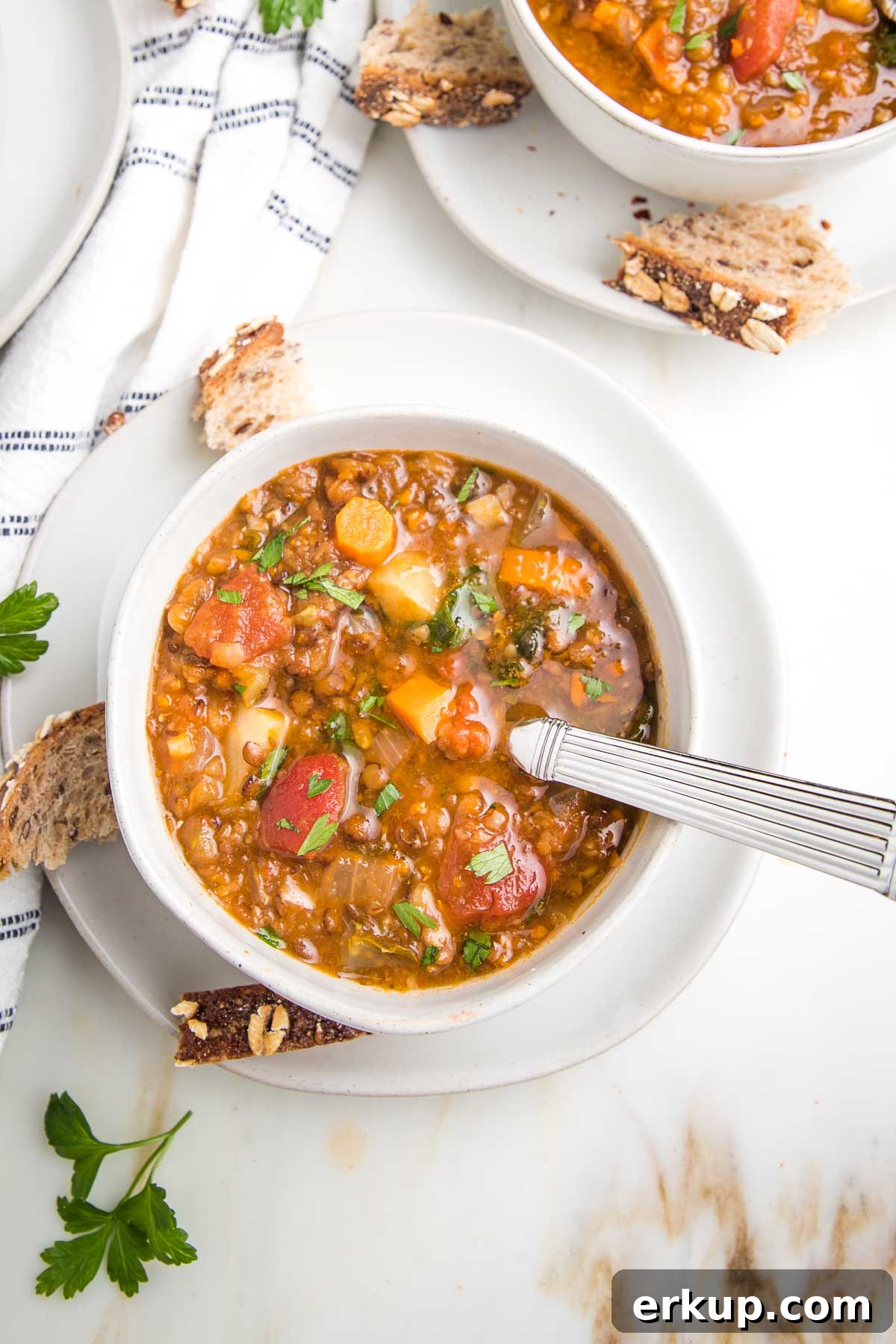 Bowl of chunky lentil soup, loaded with carrots, tomatoes, parsnips and celery in a white bowl. You can see part of another bowl in the distance. There are chunks of crusty bread and fresh parsley, plus a white dish towel with navy stripes.