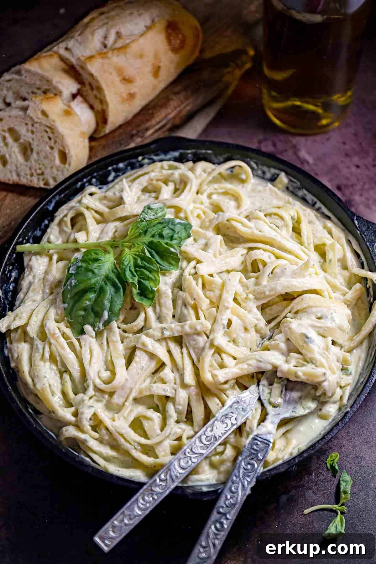 Overhead shot of pesto pasta garnished with basil in a saucepan with silverware on the side and bread in the background.