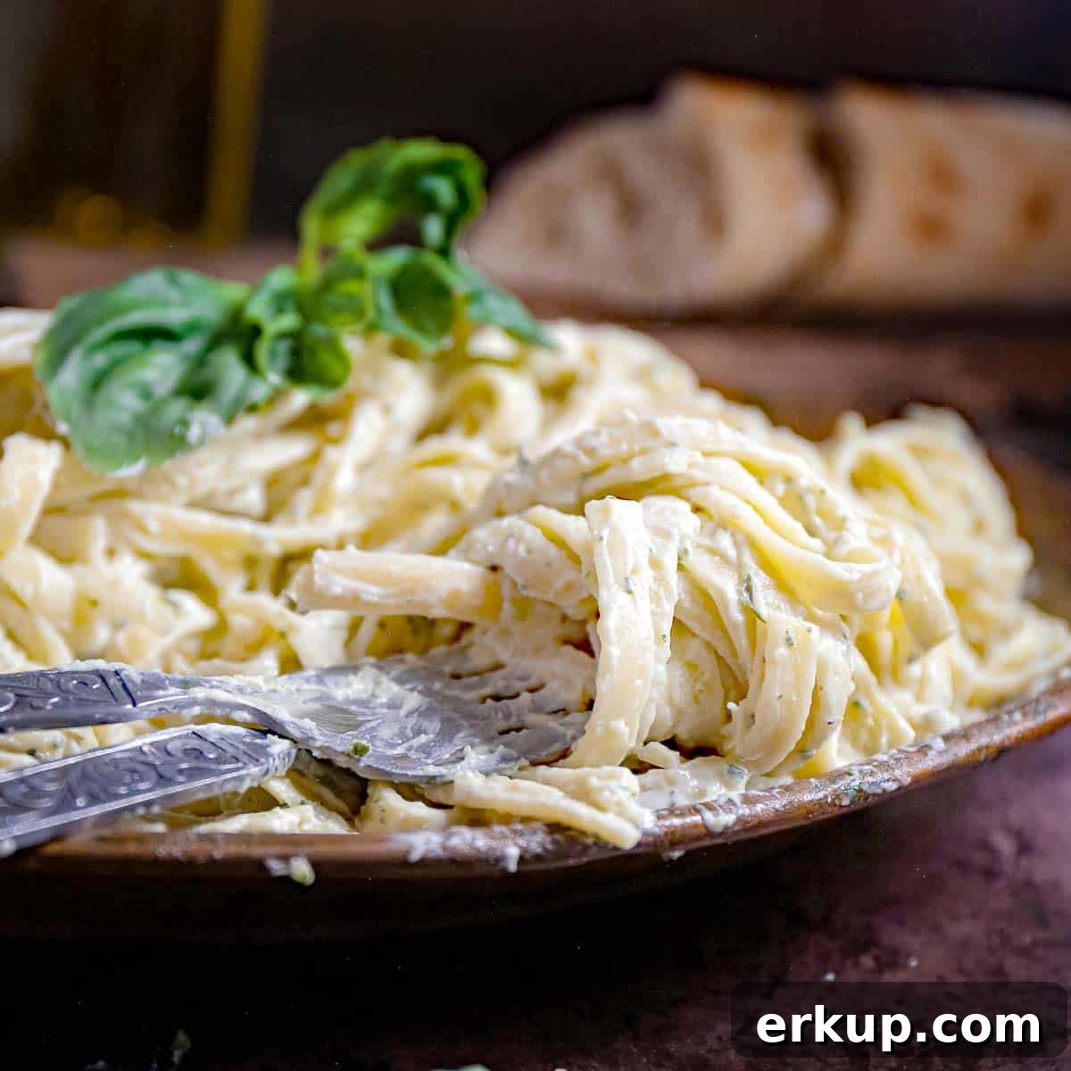 Side shot of pesto pasta garnished with basil in a wooden bowl with silverware on the side and sliced bread in the background.