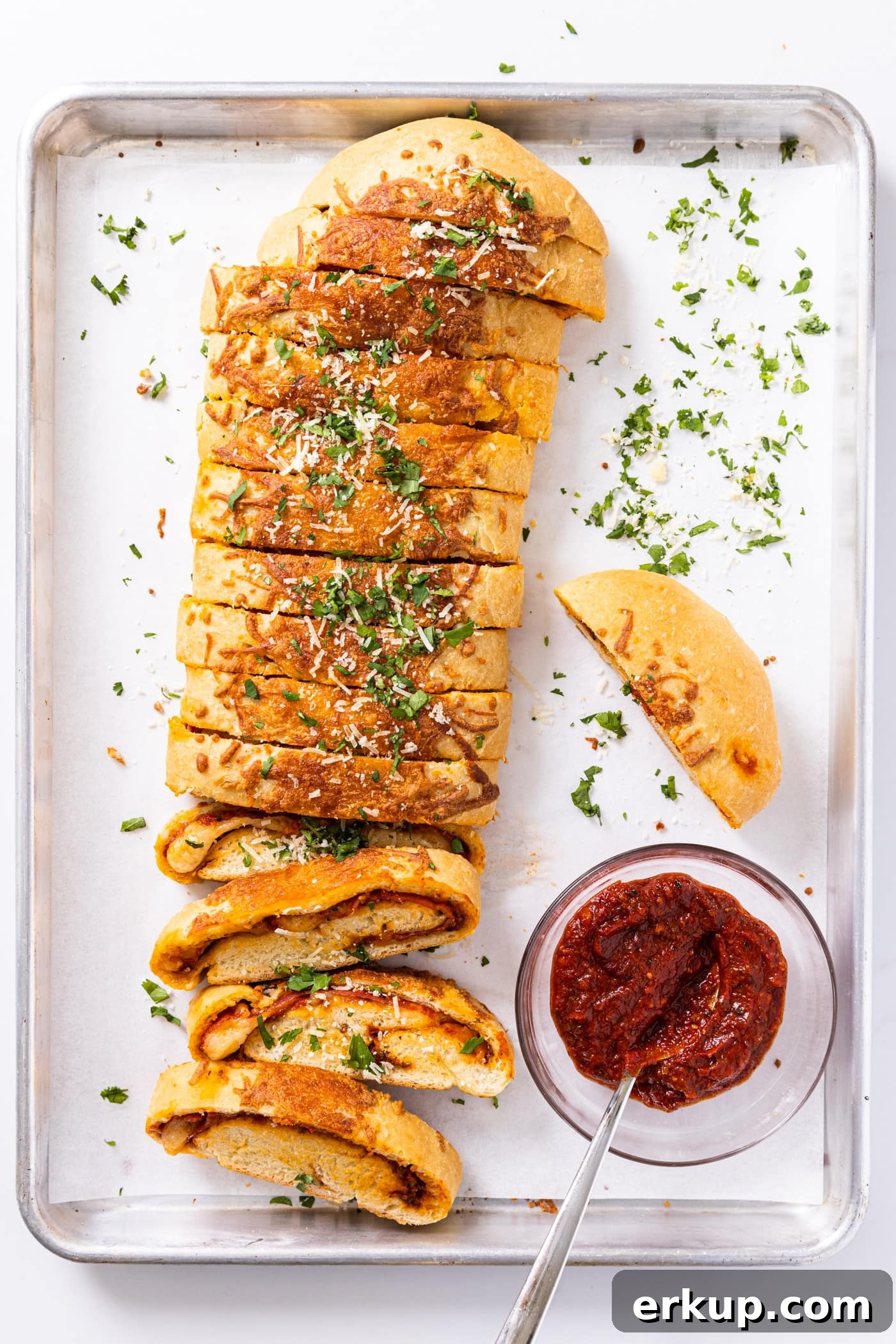 Homemade Stromboli Recipe: Crispy Cheesy Italian Roll 3 Top down view of metal baking sheet with long sliced stromboli sitting on white parchment paper with small bowl of red tomato sauce
