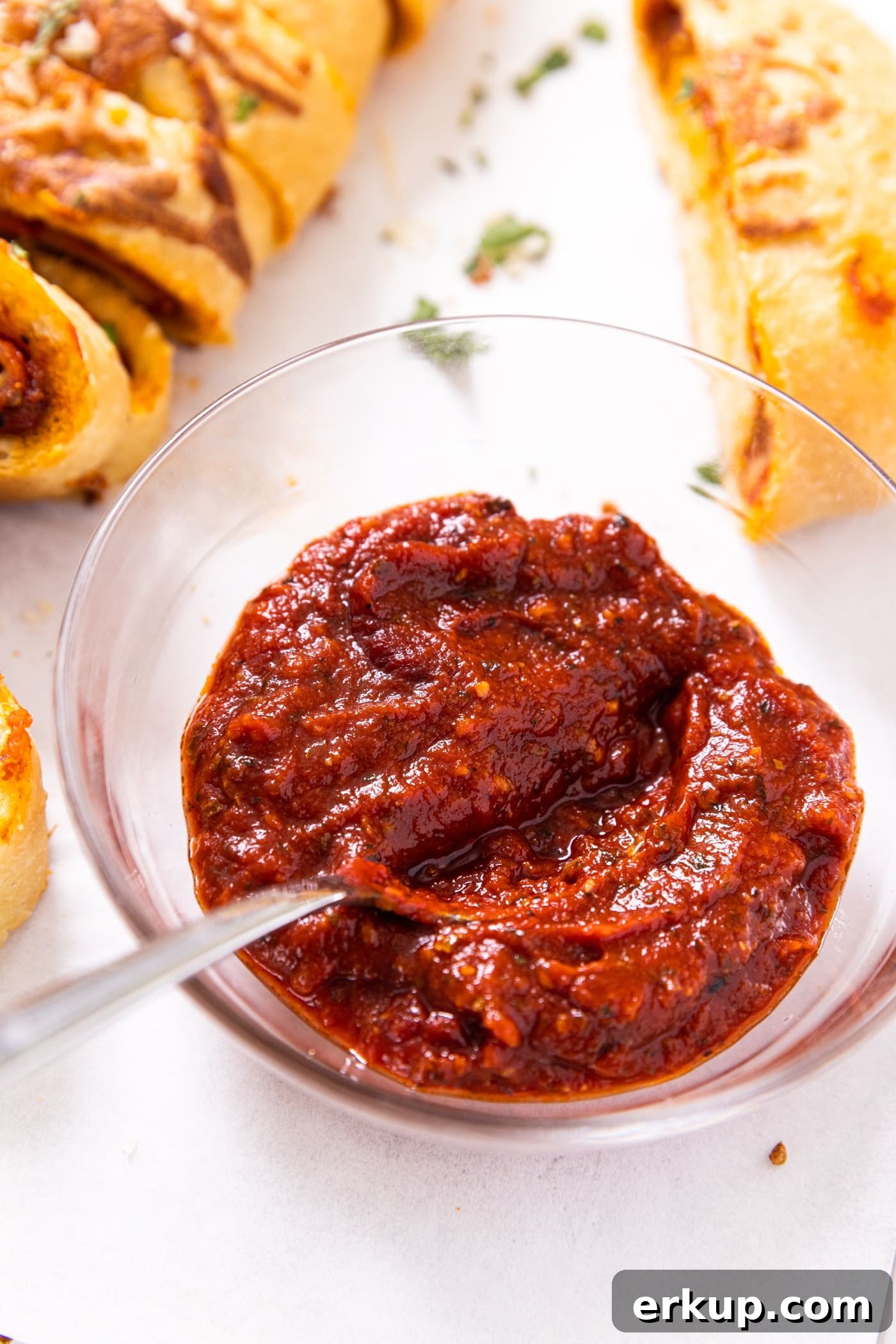 Homemade Stromboli Recipe: Crispy Cheesy Italian Roll 5 Small glass bowl filled with red tomato sauce with spoon in sauce sitting on white piece of parchment paper with slices of stromboli beside