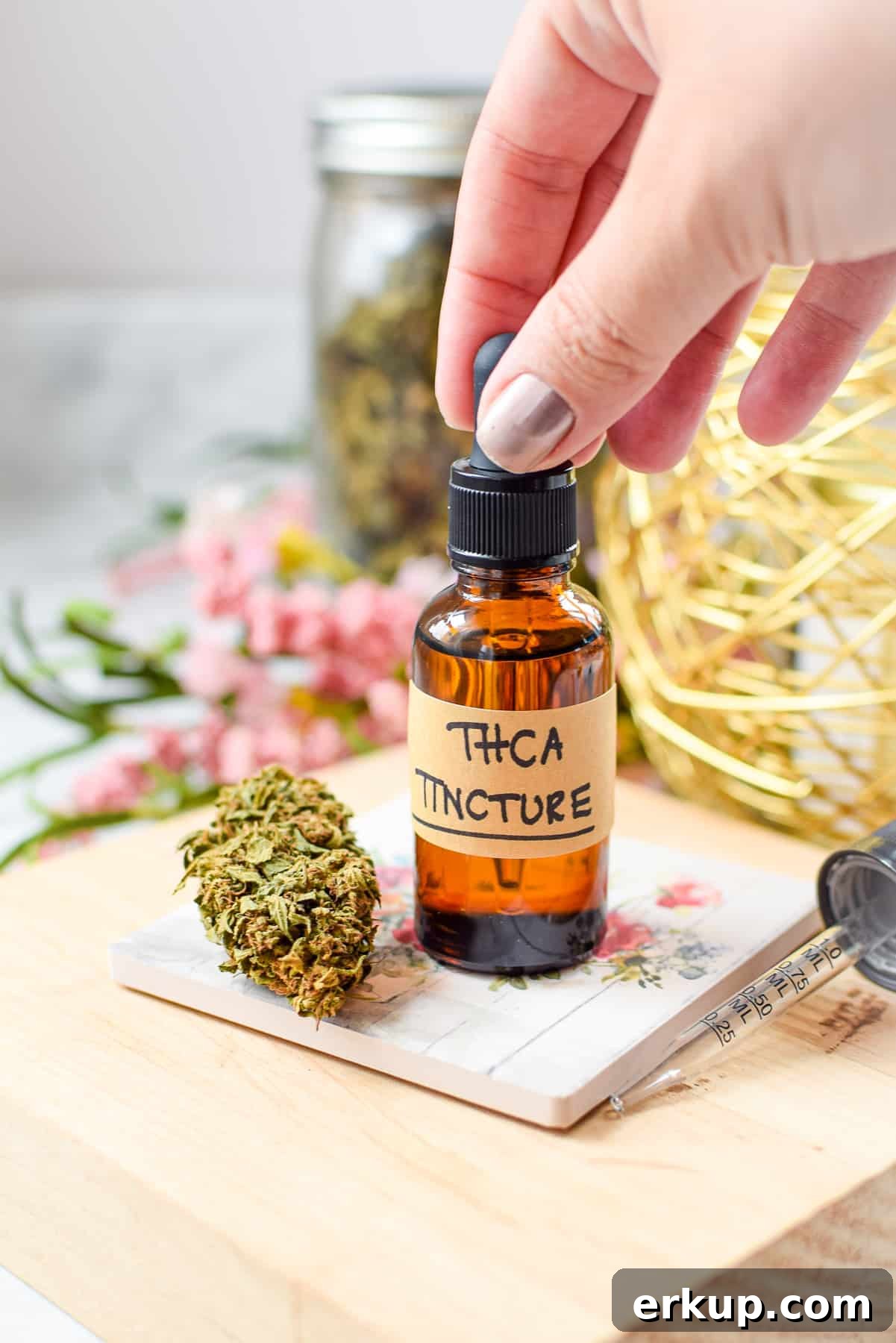 How to Make a THCA Tincture at Home 6 A white countertop with a brown tincture bottle labeled THCA tincture with a cannabis flower bud next to it with a hand gripping the top of the tincture dropper