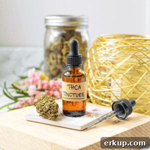 A white countertop with a brown tincture bottle labeled THCA tincture with a cannabis flower bud next to it with a hand gripping the top of the tincture dropper