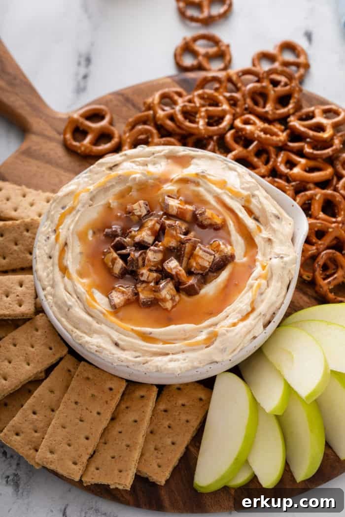 Bowl of snickers dip on a wooden board surrounded by graham crackers, sliced apples, and pretzel twists.