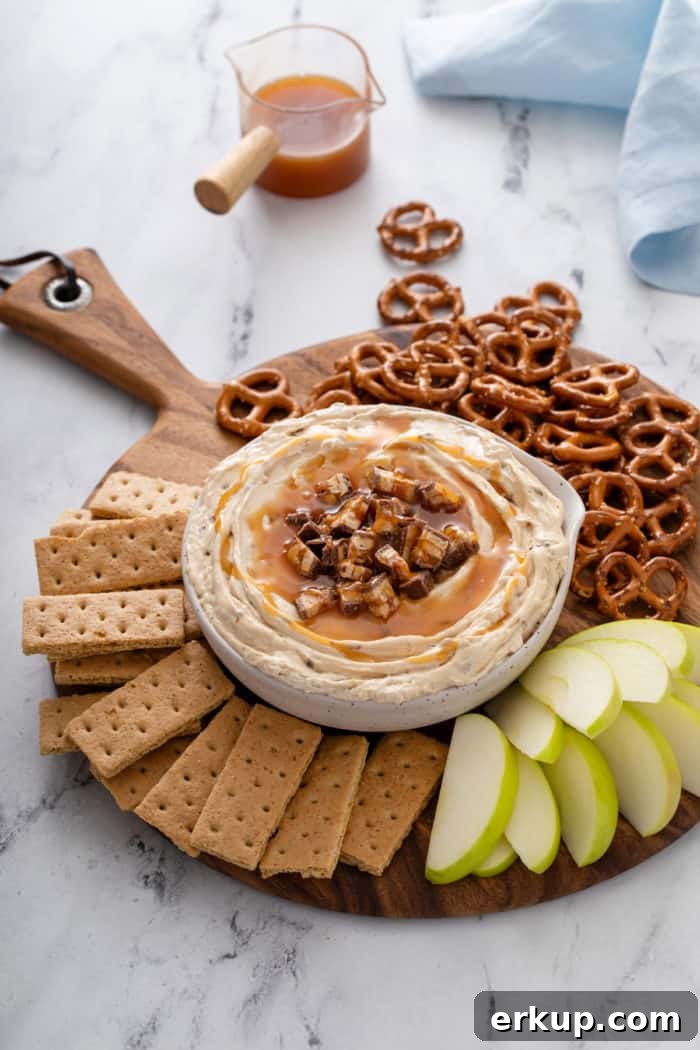 Wooden board arranged with a bowl of snickers dip, graham crackers, apples, and pretzels.