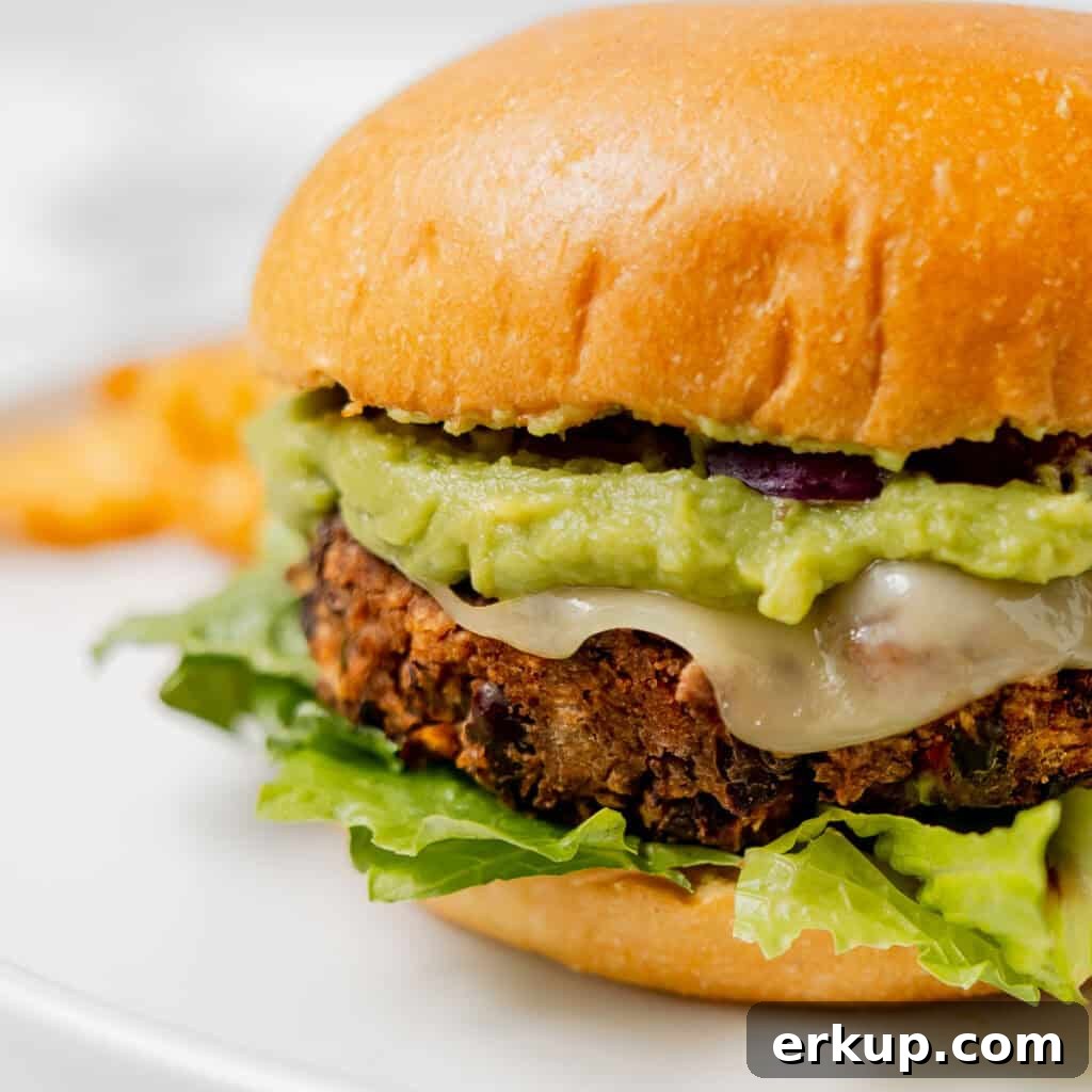 Close-up of a black bean burger with lettuce, melted cheese, and guacamole in a toasted bun, served on a white plate.