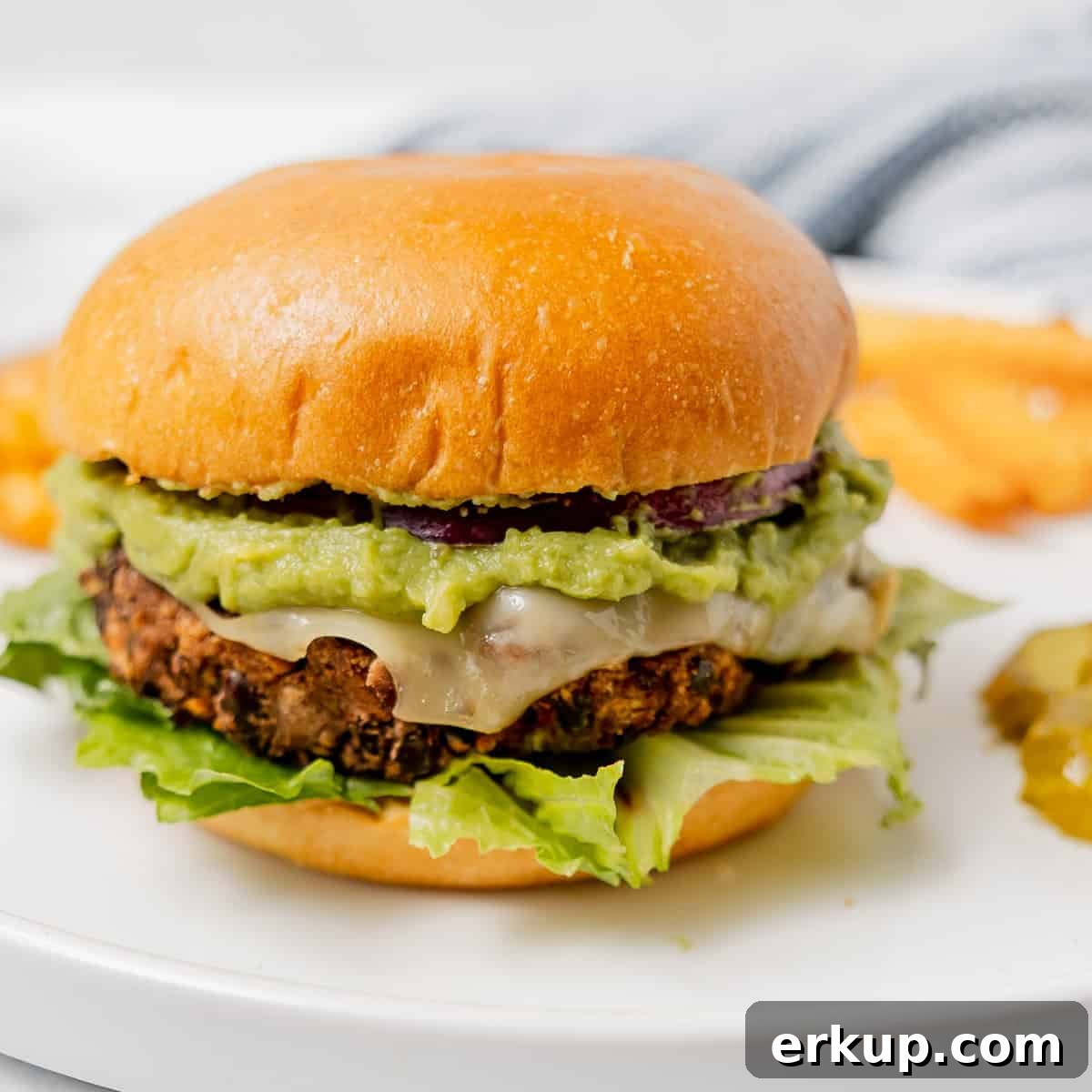 A veggie burger with lettuce, melted cheese, guacamole, and onion on a bun, served on a white plate with pickles and fries in the background.