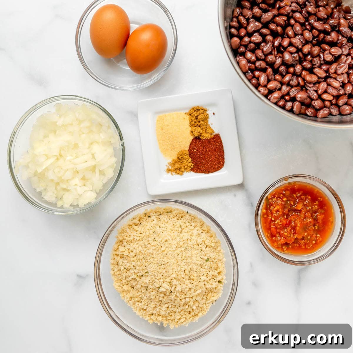 Overhead view of ingredients in bowls: black beans, eggs, chopped onions, panko breadcrumbs, salsa, and various ground spices on a small plate.