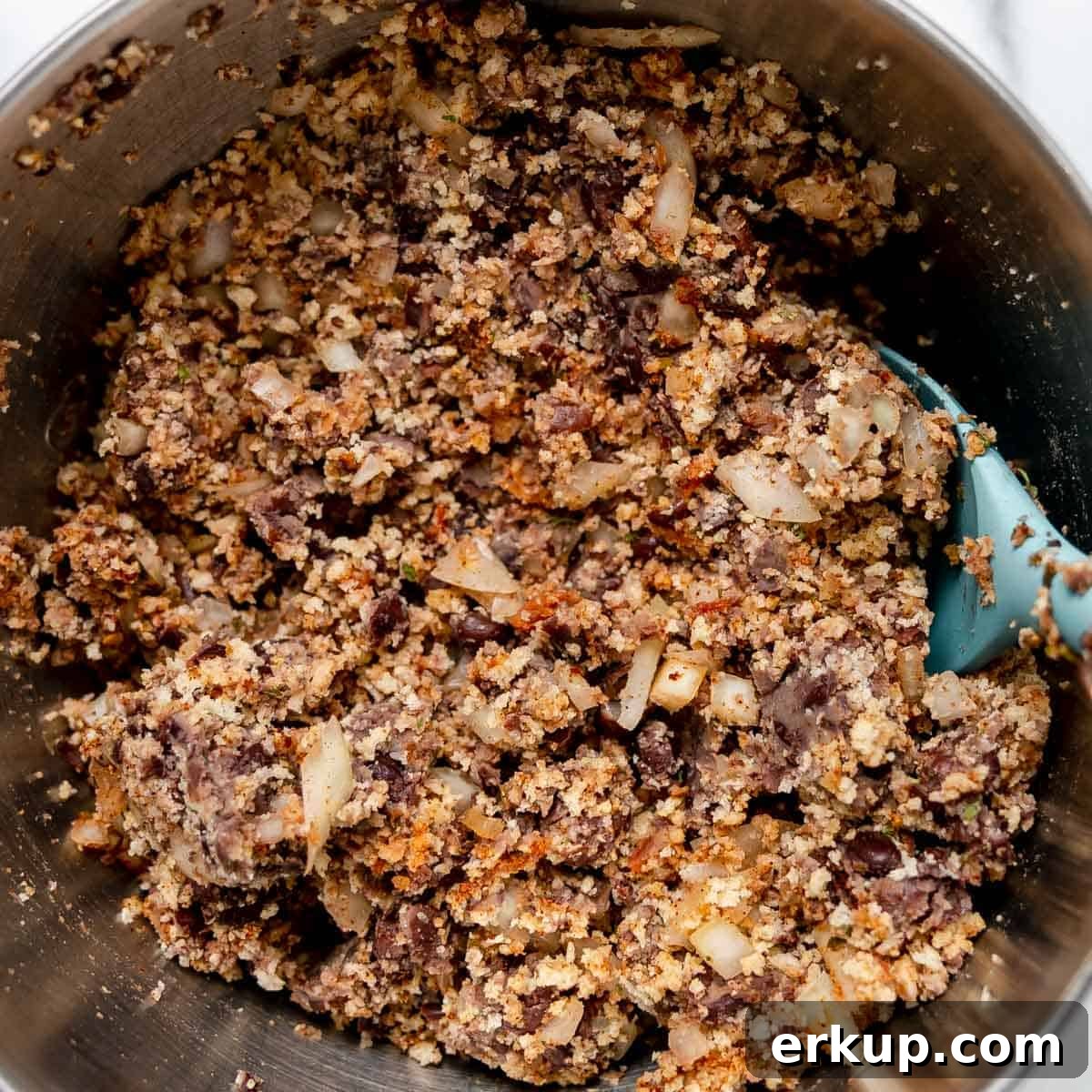 A metal bowl containing a mixture of chopped onions, beans, and breadcrumbs, with a blue spatula partially visible on the right side.