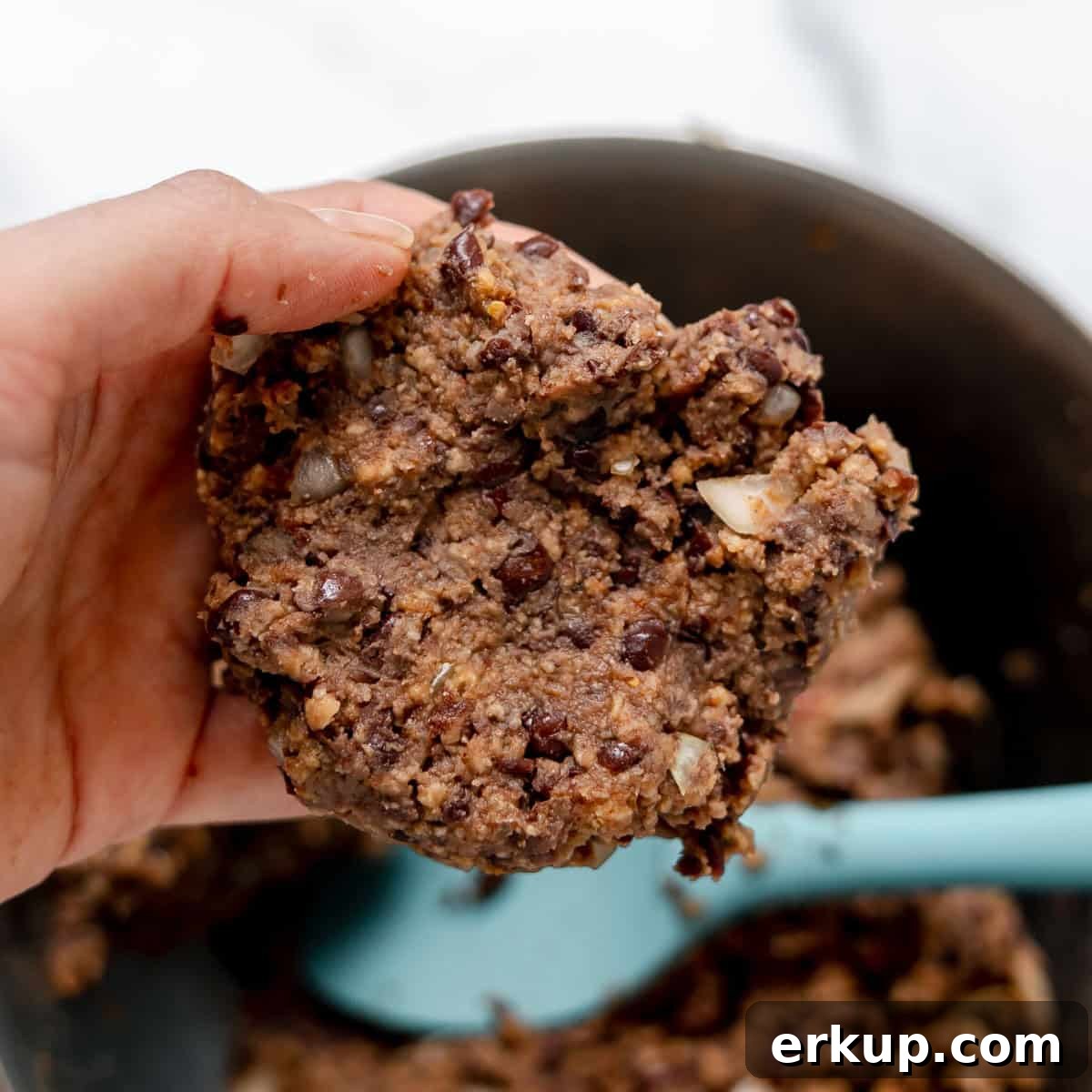 A hand holding a portion of chunky, uncooked black bean burger mixture above a bowl with a blue spatula inside.