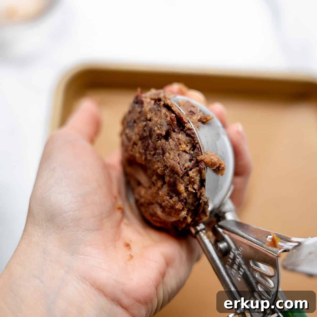 A hand holds a cookie scoop filled with cookie dough over a parchment-lined baking sheet.
