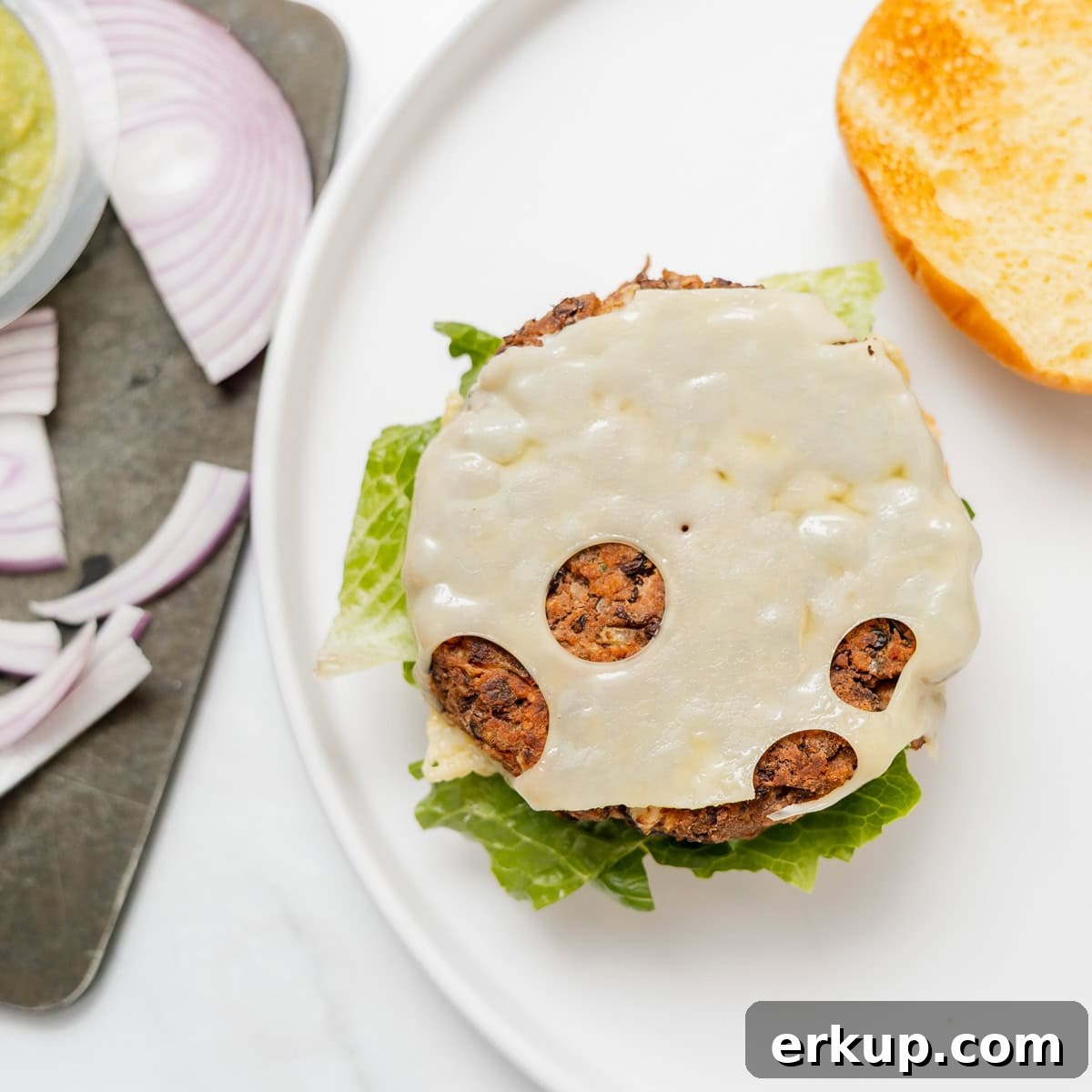 A veggie burger patty with melted Swiss cheese and lettuce on a bun, with sliced red onions and a condiment on the side.