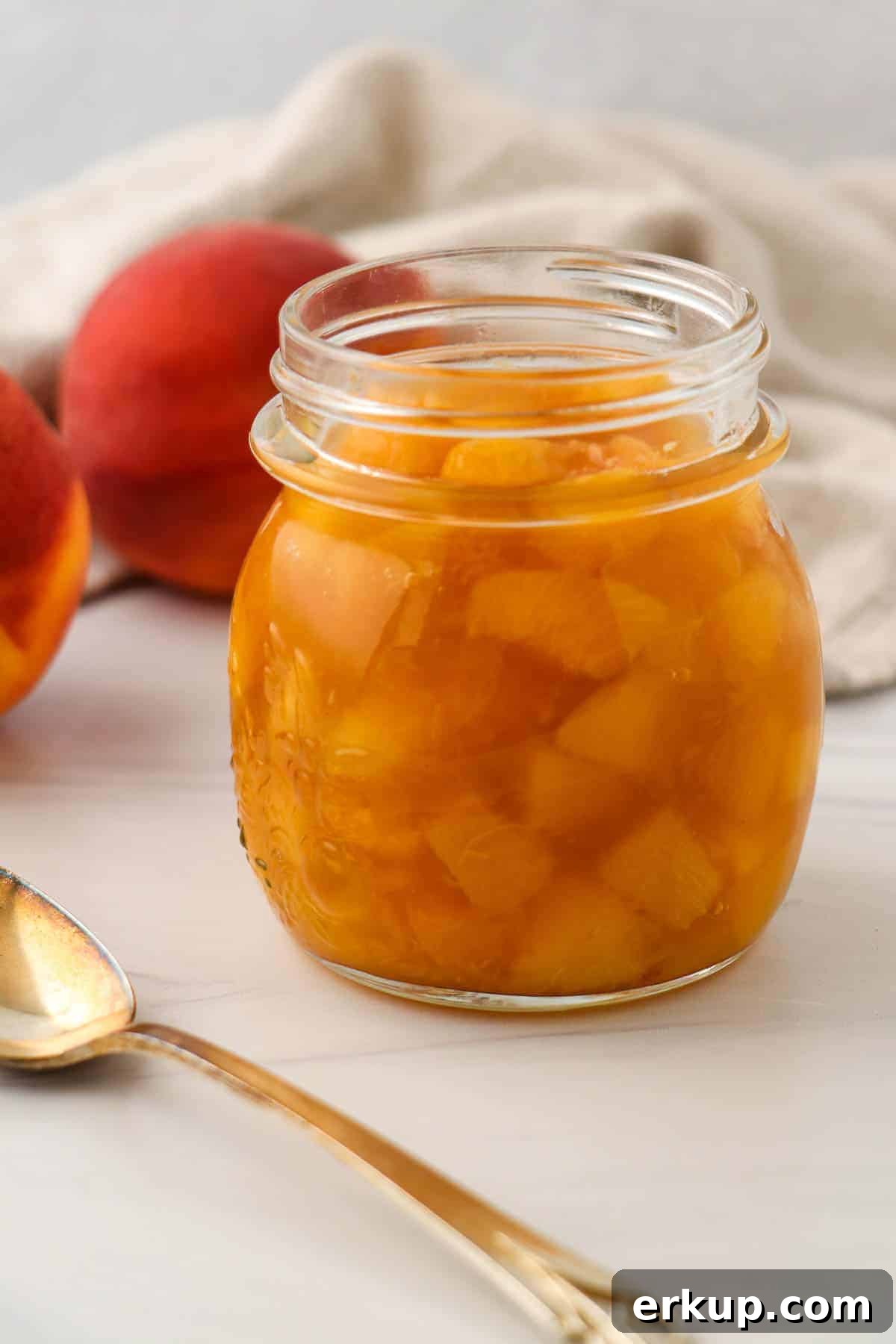 Peach compote in a jar next to a spoon and fresh peaches.