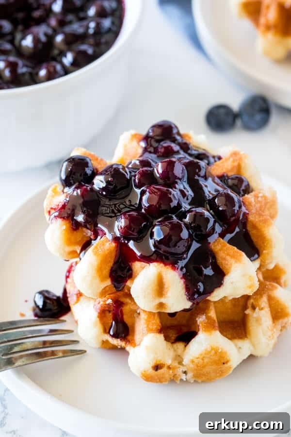 A stack of 2 small waffles topped with blueberry syrup with a bowl of blueberry sauce in the background.