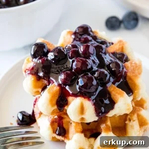 A stack of 2 small waffles topped with blueberry syrup with a bowl of blueberry sauce in the background.
