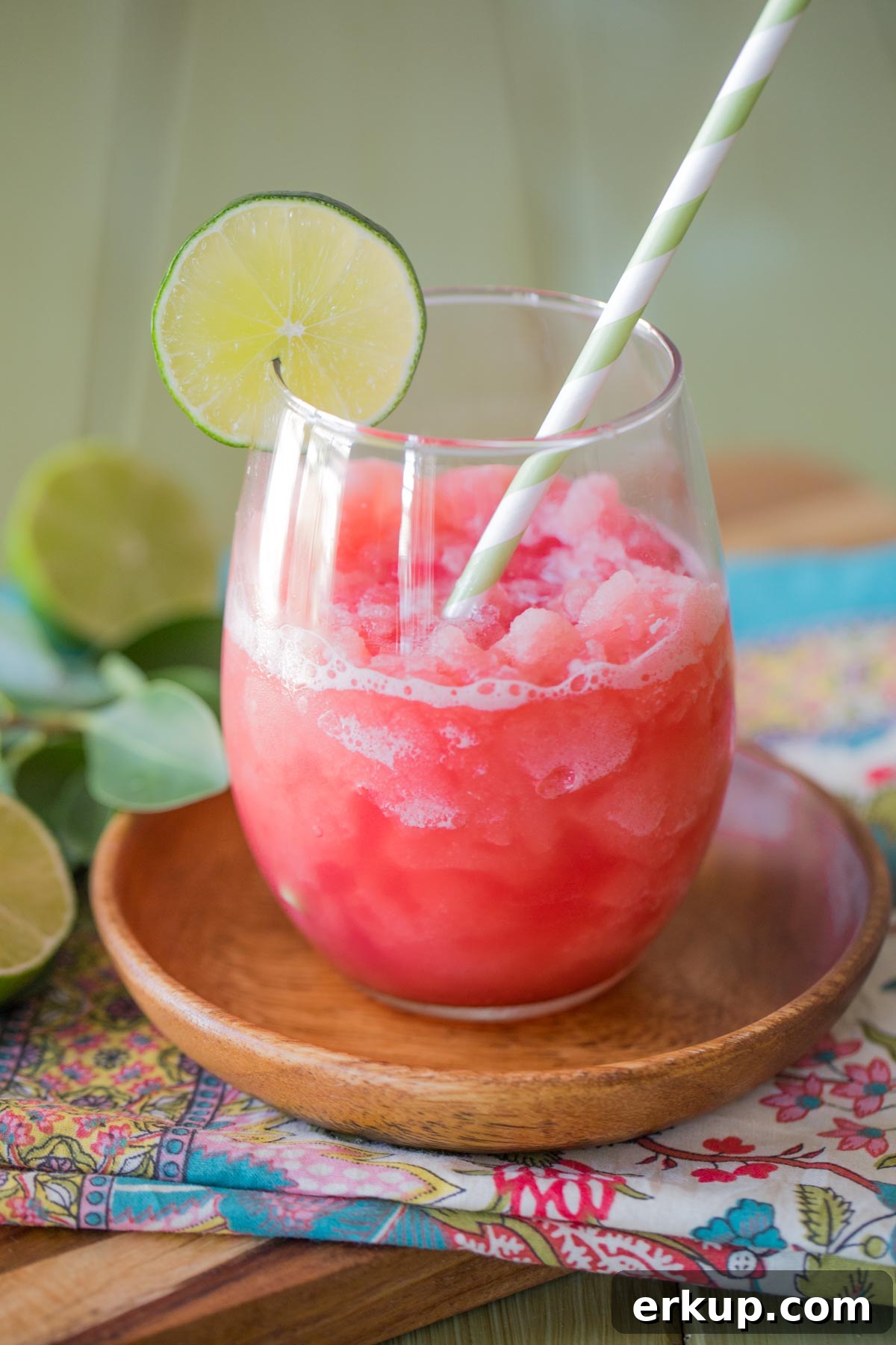 Coconut Lime Watermelon Slushie in a glass tumbler with a straw and a slice of lime on the rim, sitting on a small wood plate.