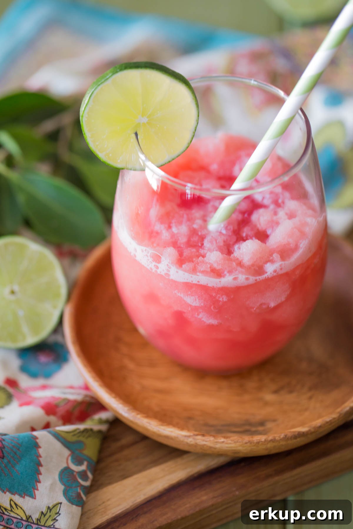 Coconut Lime Watermelon Slushie in a glass tumbler with a straw and a slice of lime on the rim, sitting on a small wood plate.