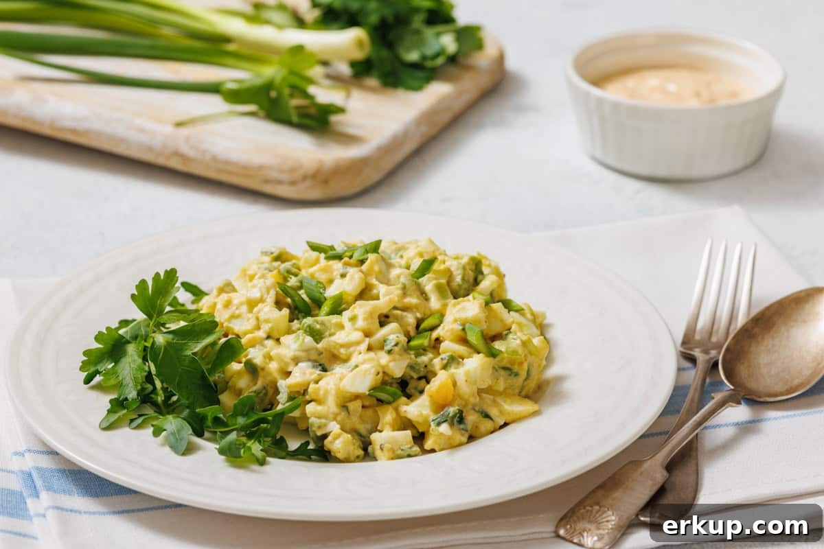 A white plate with avocado egg salad, garnished with herbs, is shown with a fork, spoon, cloth, and a small bowl of sauce.