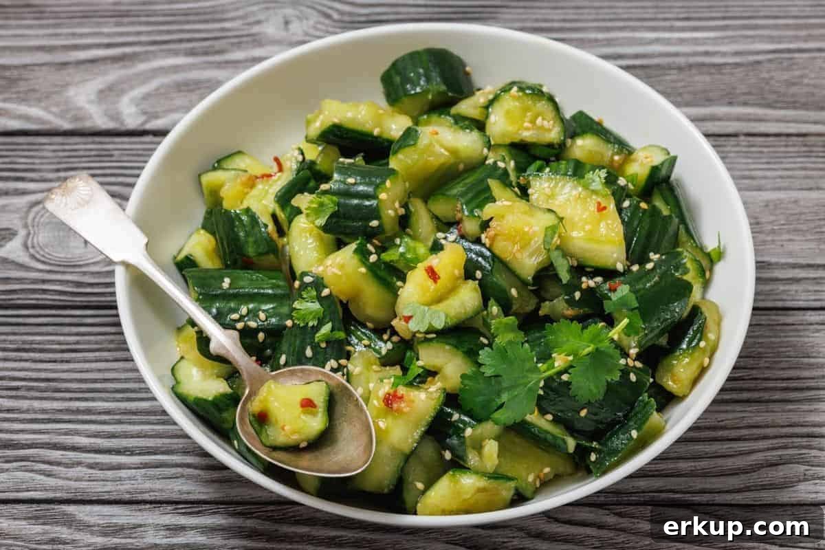 A bowl of smashed cucumber salad garnished with cilantro, sesame seeds, and chili flakes, with a spoon on the side, set on a wooden surface.