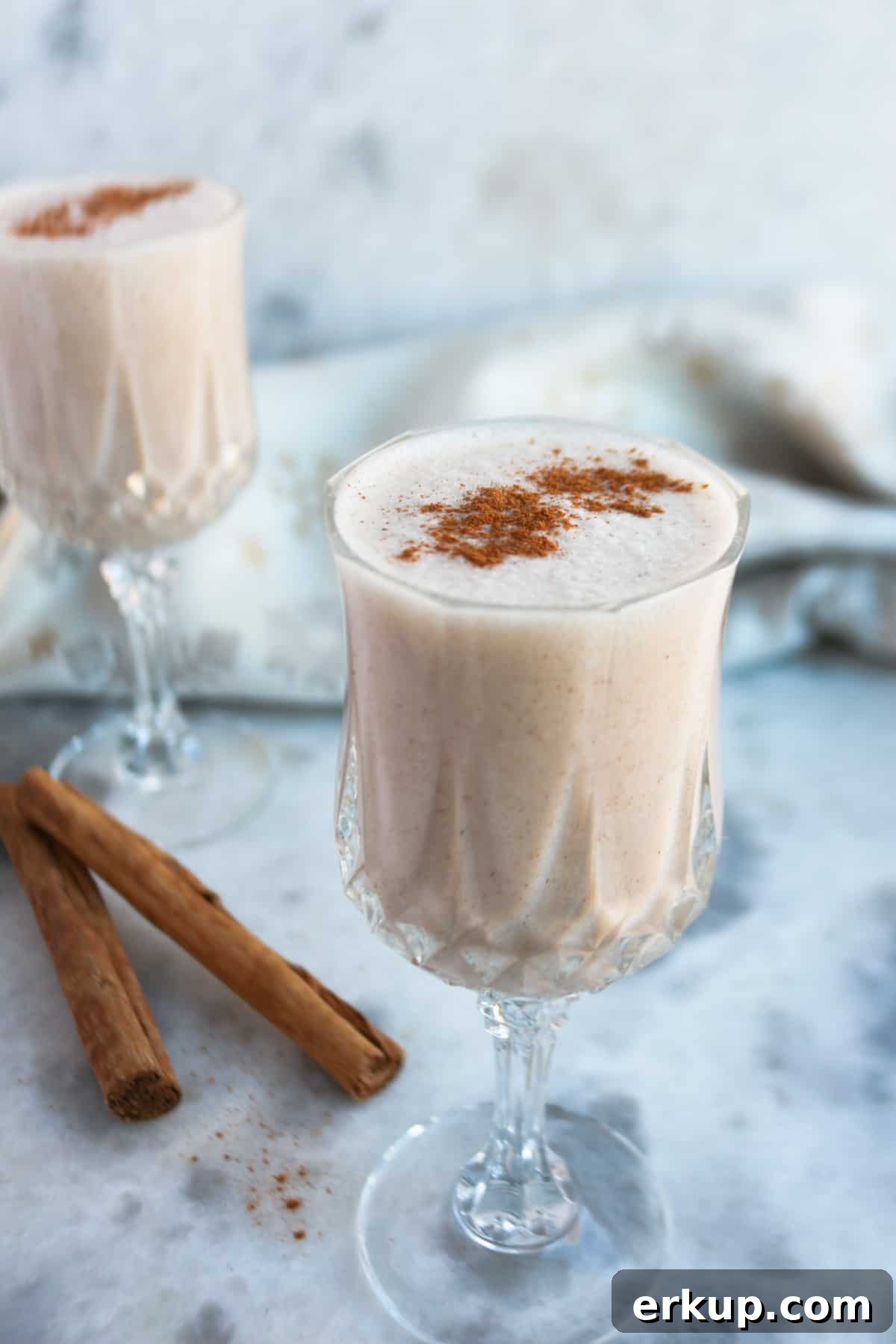 Creamy Vegan Coquito Recipe for the Holidays 2 Two clear and small wine glasses filled with coquito with cinnamon sticks beside them.